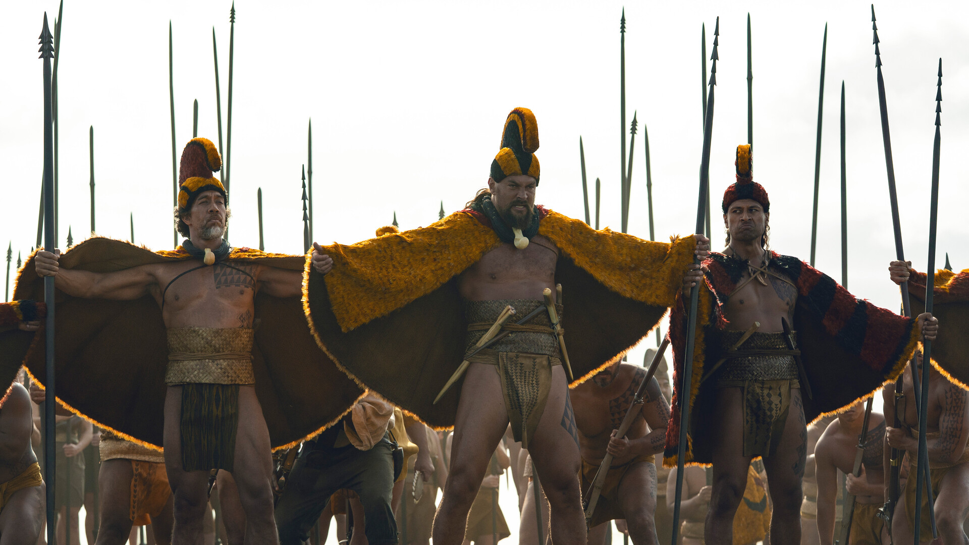 Jason Momoa between two men standing in front of a war party all dressed in traditional Hawaiian battle garb